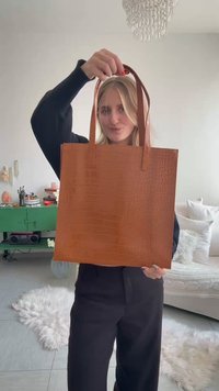 Jeune femme tenant un grand sac cabas en cuir marron à motif crocodile dans un salon lumineux et moderne.