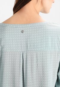 Blouse vert clair avec un motif floral, encolure ronde et un petit bouton en métal à l'arrière. Présente un design plissé pour plus de texture.