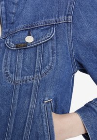 Denim jacket in blue with a buttoned chest pocket, contrasting stitching, and side pockets. Smooth texture, classic design, and branded hardware.