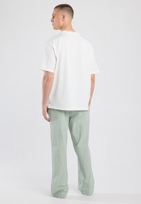 Man staat, kijkt naar links met het hoofd gedraaid, in een eenvoudig wit T-shirt en groen-witte verticaal gestreepte wijde broek.