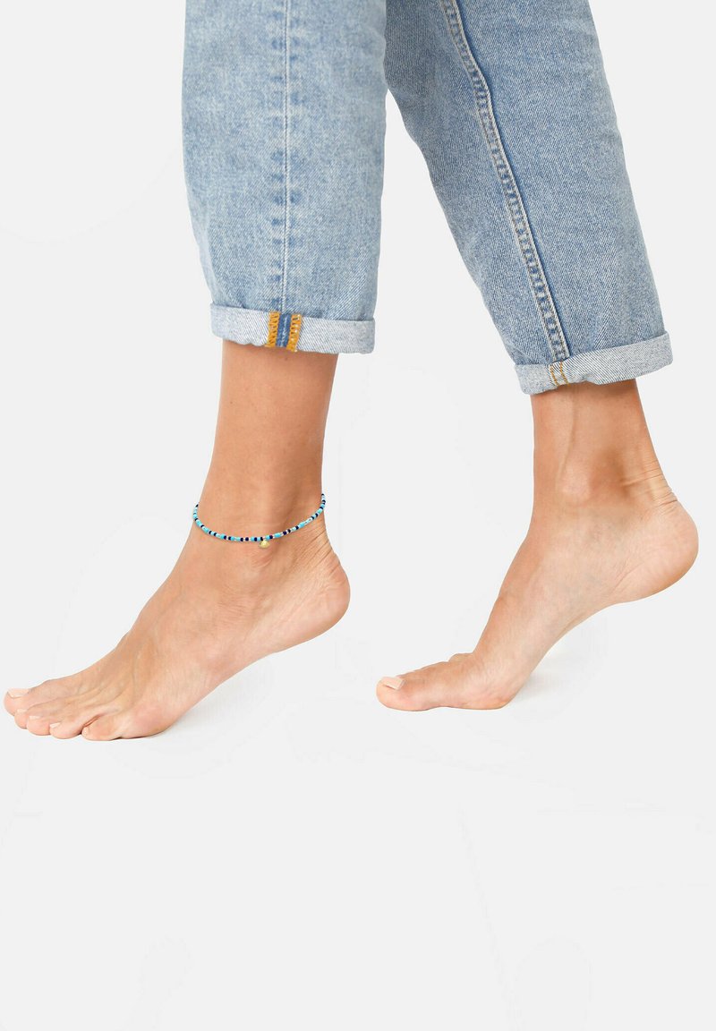 Enkelband met meerkleurige kraaltjes, met tinten blauw en kleine accenten, gedragen op een blote voet naast opgerolde spijkerbroek.