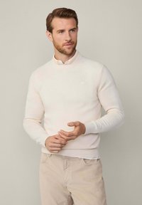 Muž s krátkými hnědými vlasy a vousy, oblečený v krémovém svetr over košilí s límečkem a béžových kalhotách, stojící proti jednobarevnému béžovému pozadí.