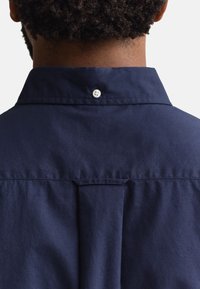 Rücken einer dunkelhäutigen Person, die ein marineblaues Hemd mit Kragen trägt, mit einem weißen Knopf an der Rückseite des Kragens und plissiertem Stoff darunter.