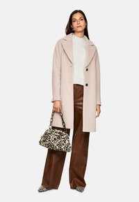 Hellrosa Mantel, grob gestrickter weißer Pullover, braune Lederhose und Handtasche mit Leopardenmuster und Kettenriemen, passende Schuhe.