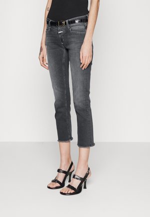 Mujer con jeans negros deslavados y recortados con dobladillos sin rematar, cinturón negro y sandalias negras de tacón alto, de pie frente a un fondo blanco.