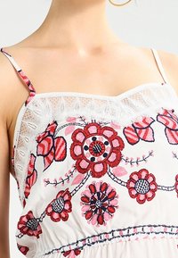 Broderad blus med flerfärgade blommönster, vit tyg, spetskant vid halsringningen och tunna justerbara axelremmar.