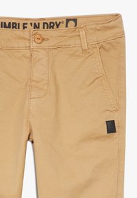 Beige puuvillahousut, joissa on nappisulku, etutaskut ja pieni musta logo-aukko sivutaskun lähellä.