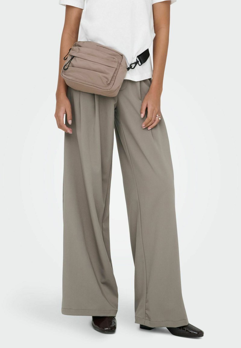 Personne portant un pantalon large beige, une chemise blanche à manches courtes, des chaussures noires et un sac bandoulière taupe avec des poches à fermeture éclair.