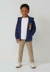 Marineblaue Zip-Hoodie mit Kängurutaschen und buntem Grafikdesign, kombiniert mit einem weißen T-Shirt und beigen Hosen, getragen mit Sneakers.