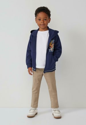 Marineblauwe zip-up hoodie met kangoeroezakken en kleurrijk grafisch ontwerp, gecombineerd met een wit T-shirt en beige broek, gedragen met sneakers.