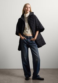 Schwarze gesteppte Cape-Jacke mit Kapuze, getragen über einem cremefarbenen Rollkragenpullover, kombiniert mit locker sitzenden blauen Jeans und schwarzem Schuhwerk.