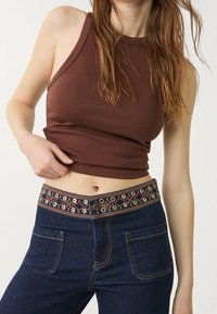 Nainen, jolla on päällään ruskea ilmanhiha crop-top ja tummansiniset farkut, joissa on brodeerattu vyö, jossa on metalliset reiät.