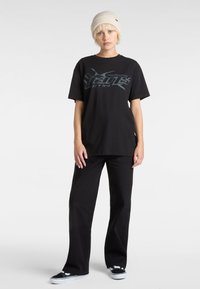Sort grafisk t-shirt med oversized pasform, prydet med grå tekstdesign. Bæres med løse sorte bukser og sorte sneakers, toppet med en cremefarvet beanie.