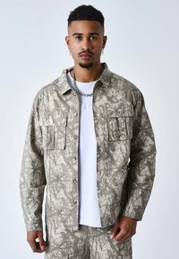 Camisa de trabajo ligera de color beige con un patrón de camuflaje, dos bolsillos en el pecho y botones en contraste. Lleva puesta una camiseta blanca debajo.