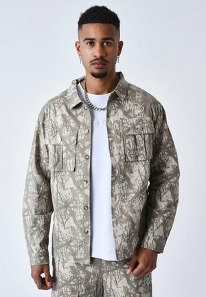 Camisa de trabajo ligera de color beige con un patrón de camuflaje, dos bolsillos en el pecho y botones en contraste. Lleva puesta una camiseta blanca debajo.
