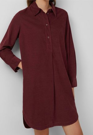 Femme portant une robe-chemise en velours côtelé rouge foncé à manches longues, avec col et une patte de boutonnage à trois boutons, main dans la poche, debout contre un mur gris.