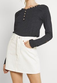 Langärmliges schwarzes Hemd mit strukturiertem Muster und Holzknöpfen am Ausschnitt, kombiniert mit einem weißen Jeans-Minirock mit Taschen.