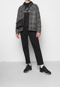 Giacca a quadri nera con toni grigi e marroni, indossata sopra una felpa nera con cappuccio, abbinata a pantaloni neri e sneakers nere, con una borsa a tracolla.