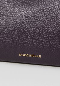 Purple leather material with a textured surface. Gold "Coccinelle" branding prominently displayed. Smooth finish.