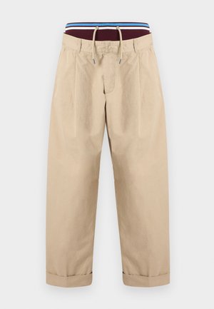 Pantalon beige avec taille à cordon, ourlet replié et détails de plis pincés. La ceinture élastique est rayée de bordeaux, bleu et blanc.