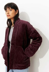 Veste en velours côtelé bordeaux avec un motif matelassé, deux poches avant et une coupe décontractée. Portée par-dessus une chemise rayée.