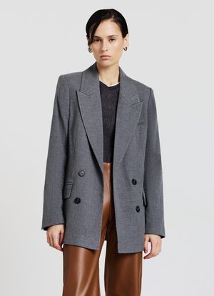 Blazer gris croisé en tissu texturé, avec des revers à pointes et des boutons noirs. Le modèle porte un pantalon en simili cuir brun en dessous.
