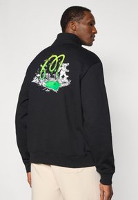 Μαύρο φούτερ με ψηλό γιακά, με έντονο πράσινο γράφημα στην πλάτη, που απεικονίζει μια αρκούδα και φυλλώματα γύρω από μια λίμνη. Απαλή υφή.