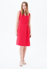 Vestido sin mangas rojo con diseño cruzado, largo hasta la rodilla y detalles plisados. La modelo lleva sandalias de tiras beige. Fondo blanco liso.