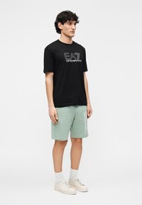Zwart katoenen T-shirt met het "EA7 Emporio Armani" logo, gecombineerd met mintgroene katoenen shorts met een gedrukt logo, en crème sneakers.