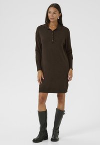 Robe en tricot marron avec un col polo et une patte de boutonnage à trois boutons. Manches longues et coupe décontractée. Portée avec des bottes en cuir noir jusqu'au genou.