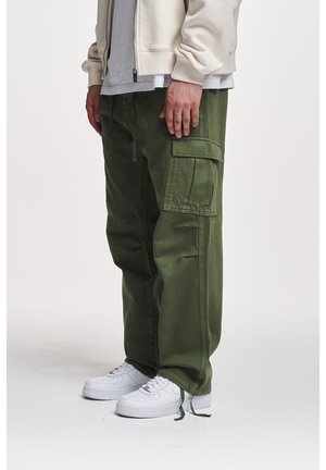 Pantalons cargo vert olive avec de nombreuses poches, fabriqués en tissu mélangé coton. Associés à des baskets blanches et une chemise gris clair en dessous.