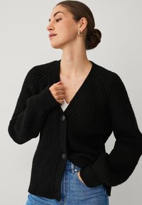 Zwarte geribbelde cardigan met een V-hals, voorzien van textuurstof, twee knopen en lange, iets oversized mouwen. Draagt over een witte top.