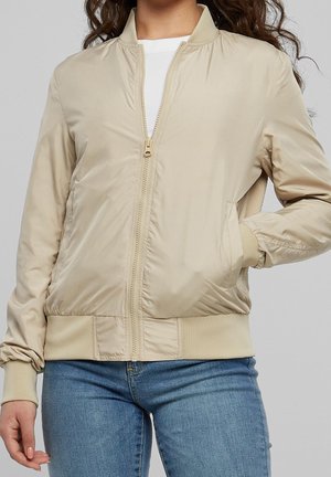 Femme portant une veste bomber beige zippée avec poignets et taille côtelés, associée à un jean bleu, debout avec une main dans la poche.