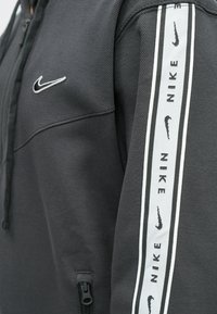 Sivi Nike dukserica s teksturiranim materijalom. Sadrži džep sa patentnim zatvaračem, mali izvezeni logo na grudima i akcent u obliku uzorka na rukavu.