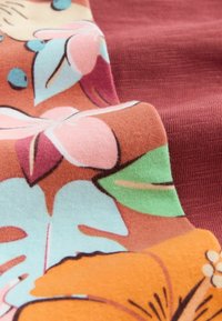 Gros plan sur un tissu doux présentant un motif floral coloré avec des pétales orange, rose, vert et bleu sur un fond rougeâtre.