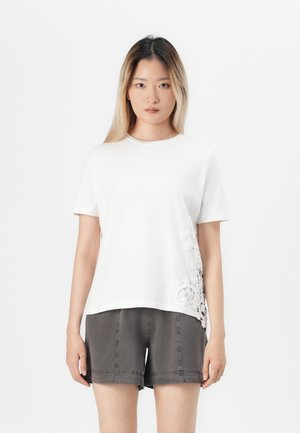 Weißes Baumwoll-T-Shirt mit einem strukturierten Spitzendetail auf der linken Seite, kurzen Ärmeln und einem klassischen Rundhalsausschnitt. Kombiniert mit grauen Shorts.