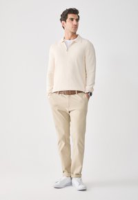 Suéter cremoso con cierre, cuello tipo solapa, combinado con chinos beige y zapatillas blancas. Diseño smart-casual, textura suave y colores neutros.
