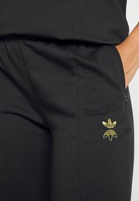 Čierne tepláky vyrobené z mäkkej tkaniny, s vyšitým zlatým logom na ľavej vrecku, elastickým pásom a funkčnými bočnými vreckami.