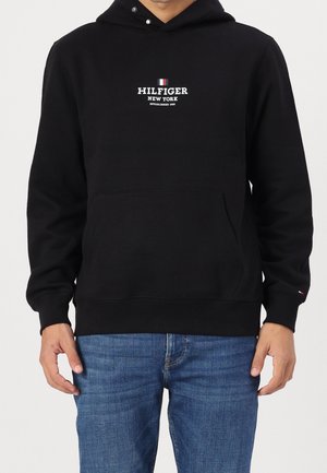 Man draagt een zwarte hoodie met een voorzak en witte tekst "Hilfiger New York", gecombineerd met blauwe spijkerbroek, staat met ontspannen handen.