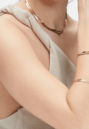 Femme portant un haut beige sans manches, un collier en or et des bracelets assortis en or au bras et au poignet.