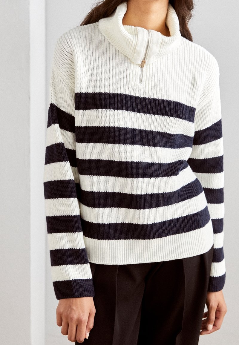 Femme portant un pull en maille blanc à rayures horizontales noires et un col à demi-fermeture éclair, associé à un pantalon noir.