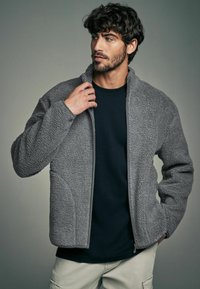 Giacca in felpa grigia con colletto alto, materiale testurizzato, chiusura con zip e tasche laterali, indossata sopra una maglietta blu scuro.