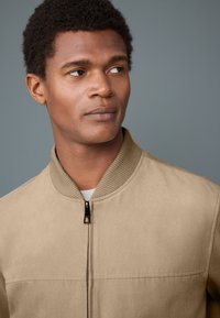Beige Bomberjacke mit strukturierter Oberfläche, geripptem Kragen und zentralem Reißverschluss. Dezente Nahtdetails entlang der Vorderpaneele.