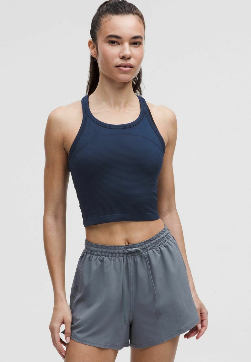 Tamno plavi rebrasti sportski crop top s tankim naramenicama, uparen sa svijetlo sivim kratkim hlačama s elastičnom pasicom, koje imaju opušteni kroj i vezicu.