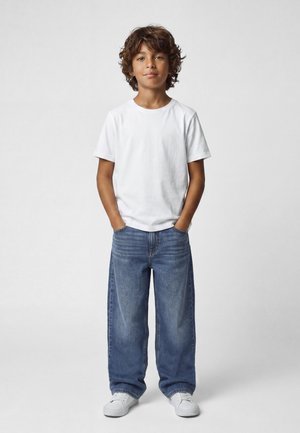 Niño de pie con las manos en los bolsillos, vistiendo una camiseta blanca, vaqueros azules holgados y zapatillas blancas sobre un fondo liso.