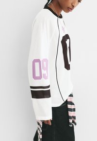 Weißes Mesh-Langarmshirt mit lila und braunen Akzenten, mit der Nummer "09" auf dem Ärmel und "0" auf der Vorderseite, schlanker Schnitt.