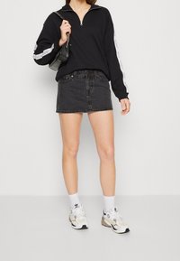 Černá zipová mikina s bílými bočními pruhy, v kombinaci s černou džínovou minisukní. Modelka má na sobě bílé tenisky s béžovými akcenty.