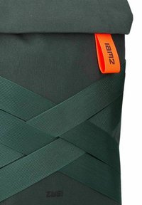 Sac à dos en tissu vert avec un haut roulé, une étiquette de logo orange et des sangles élastiques croisées pour la fermeture. Matériaux texturés et durables.