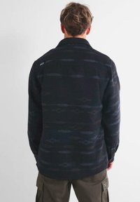 Hombre con cabello corto y ondulado visto desde atrás, vistiendo una camisa de manga larga de patrón oscuro y pantalones cargo oliva, contra un fondo blanco liso.