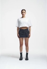 T-shirt blanc cropped avec une coupe décontractée ; mini jupe en denim noir avec deux poches avant ; bottes de combat noires à lacets. Fond uni.